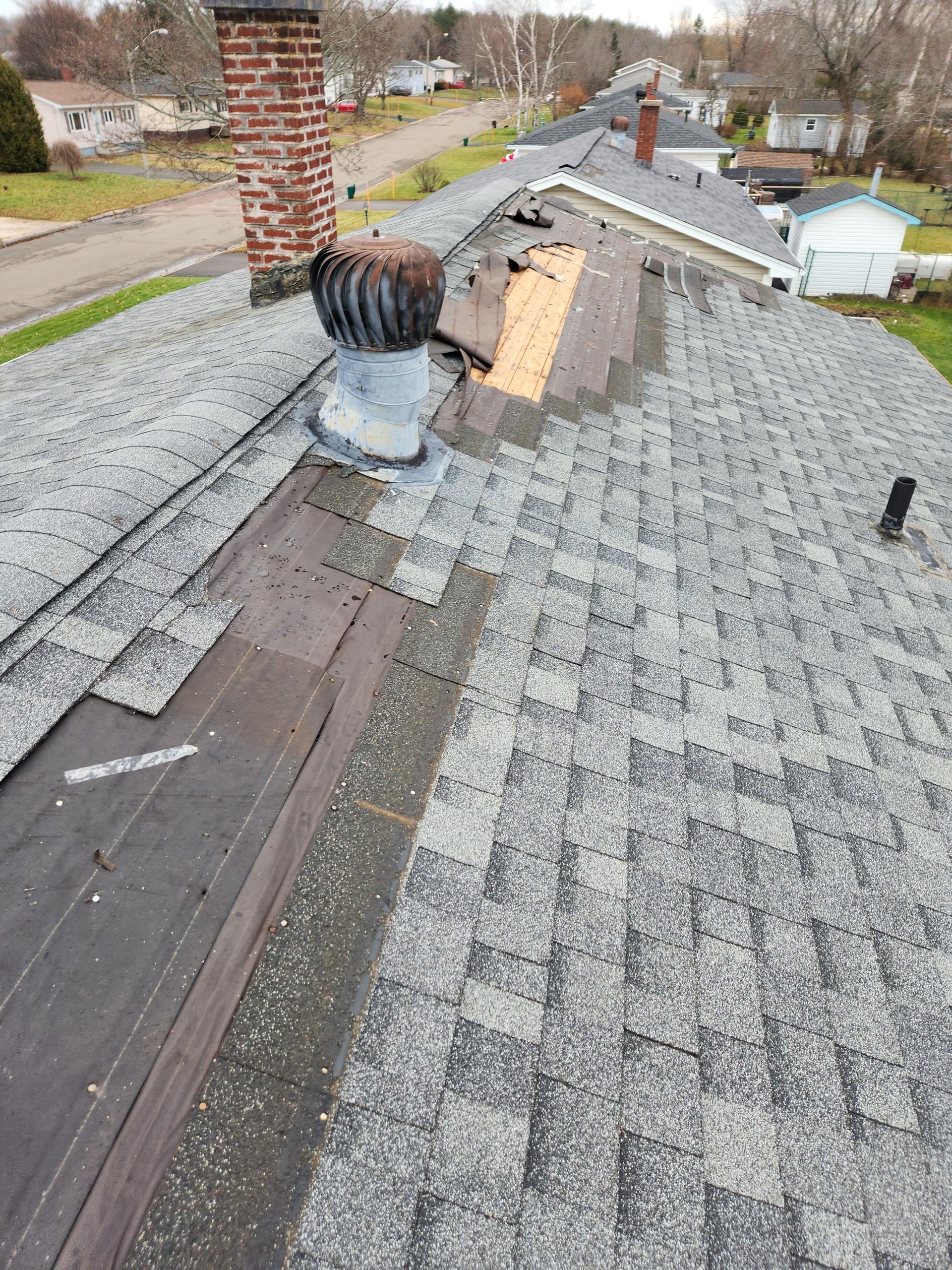 A damaged shingle roof with missing sections, exposed wooden decking, and a metal roof vent near a brick chimney.