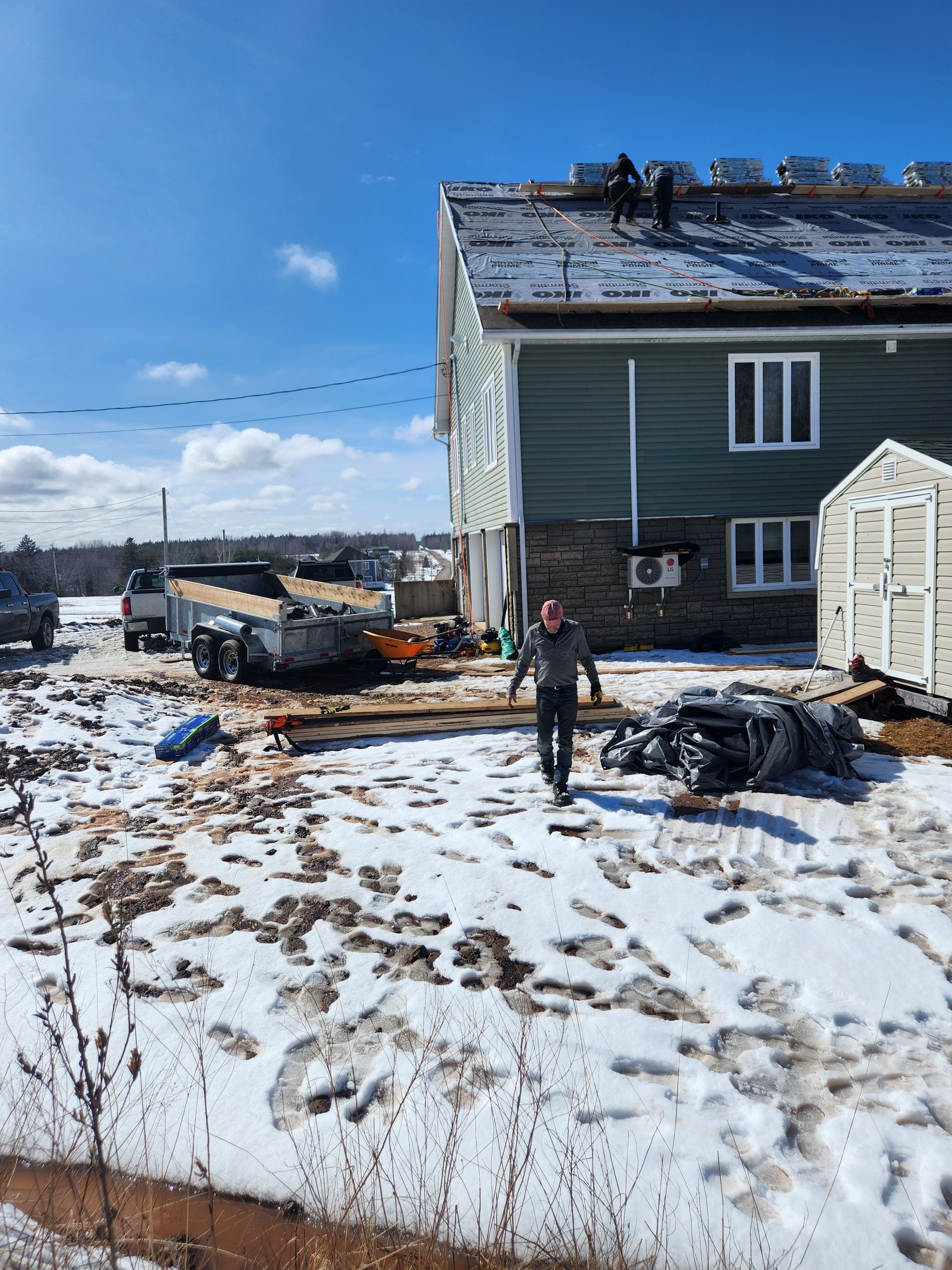 A person walks on snow near a green house where workers are replacing the roof and a trailer is parked nearby.