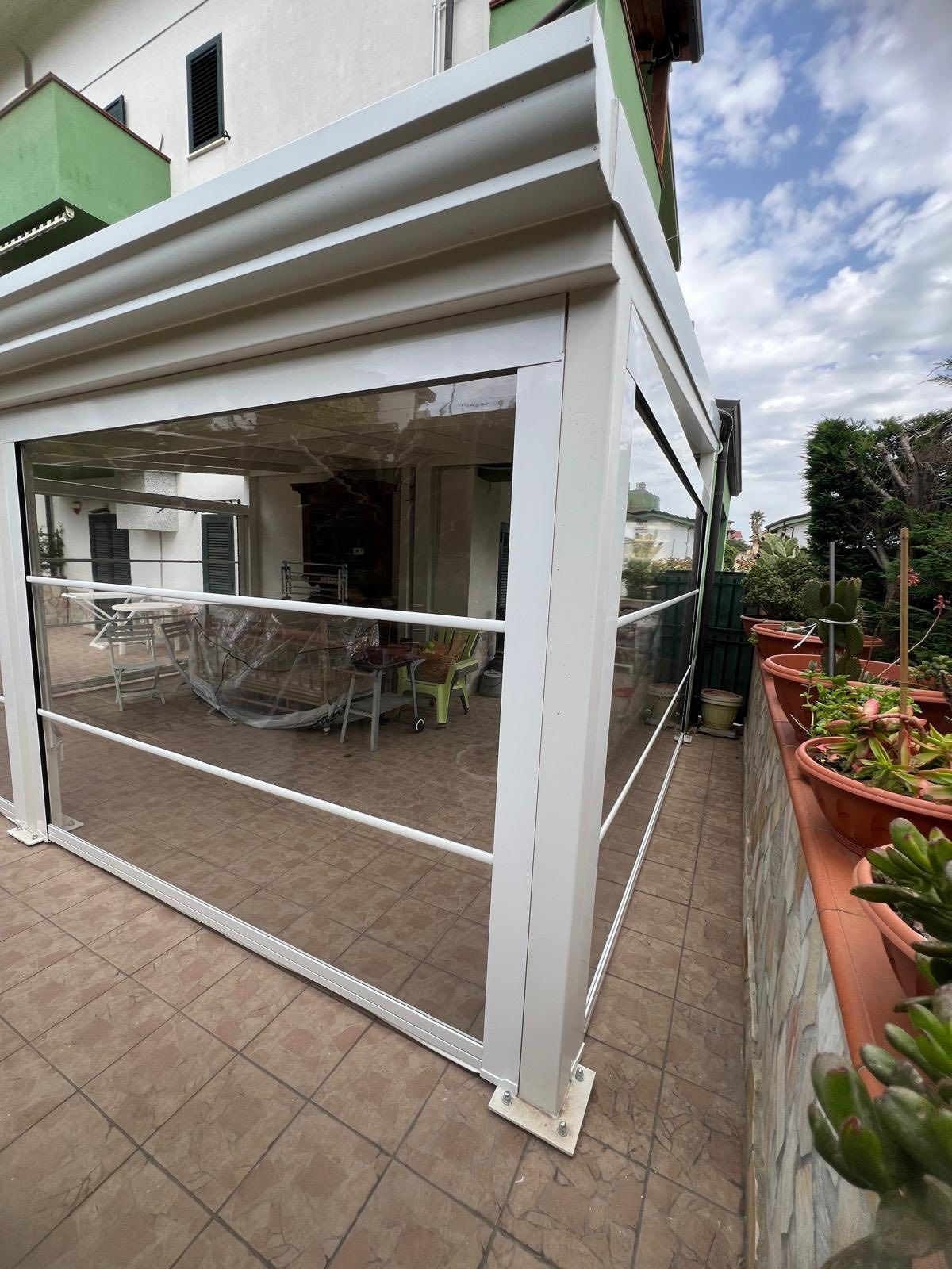 Un pergolato bianco con pareti in vetro trasparente si trova su un patio accanto a una casa.