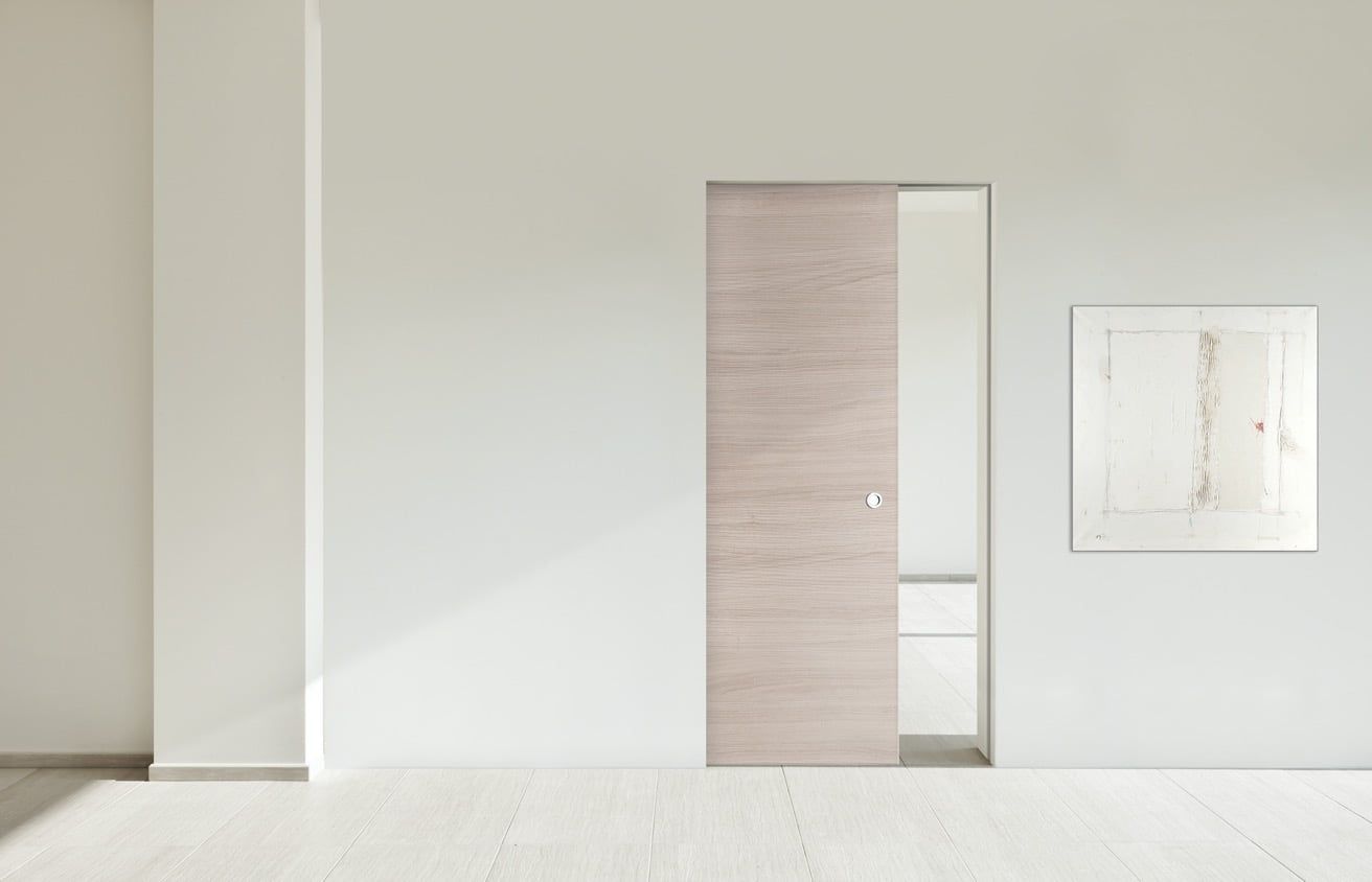 Stanza minimalista bianca con porta in legno chiaro e piccolo pannello a parete.