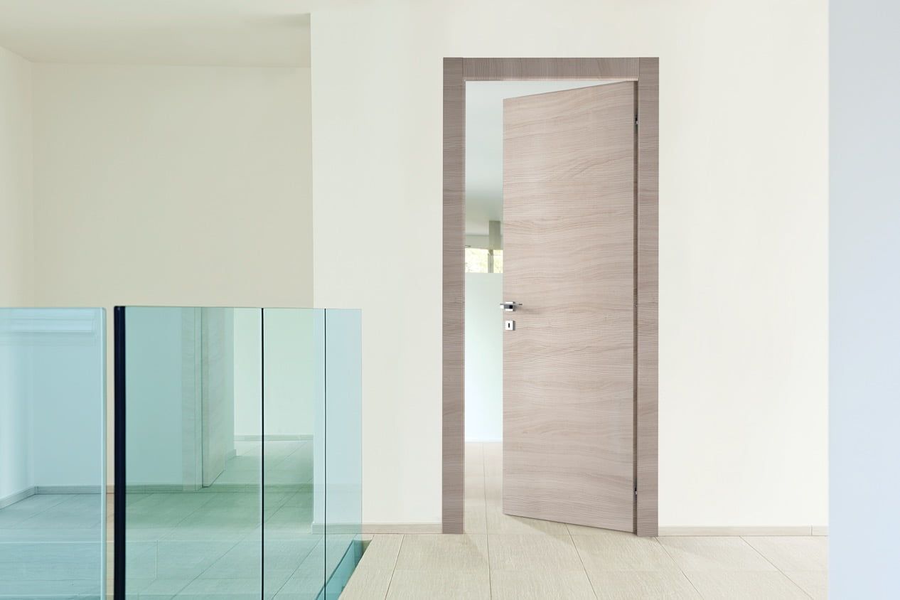 Interno minimalista con porta in legno parzialmente aperta e ringhiera in vetro blu lungo un corridoio luminoso.
