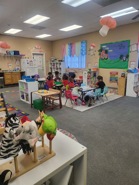 Children in a colorful classroom; several seated at tables, engaging in activities. Decorated room with puppets.