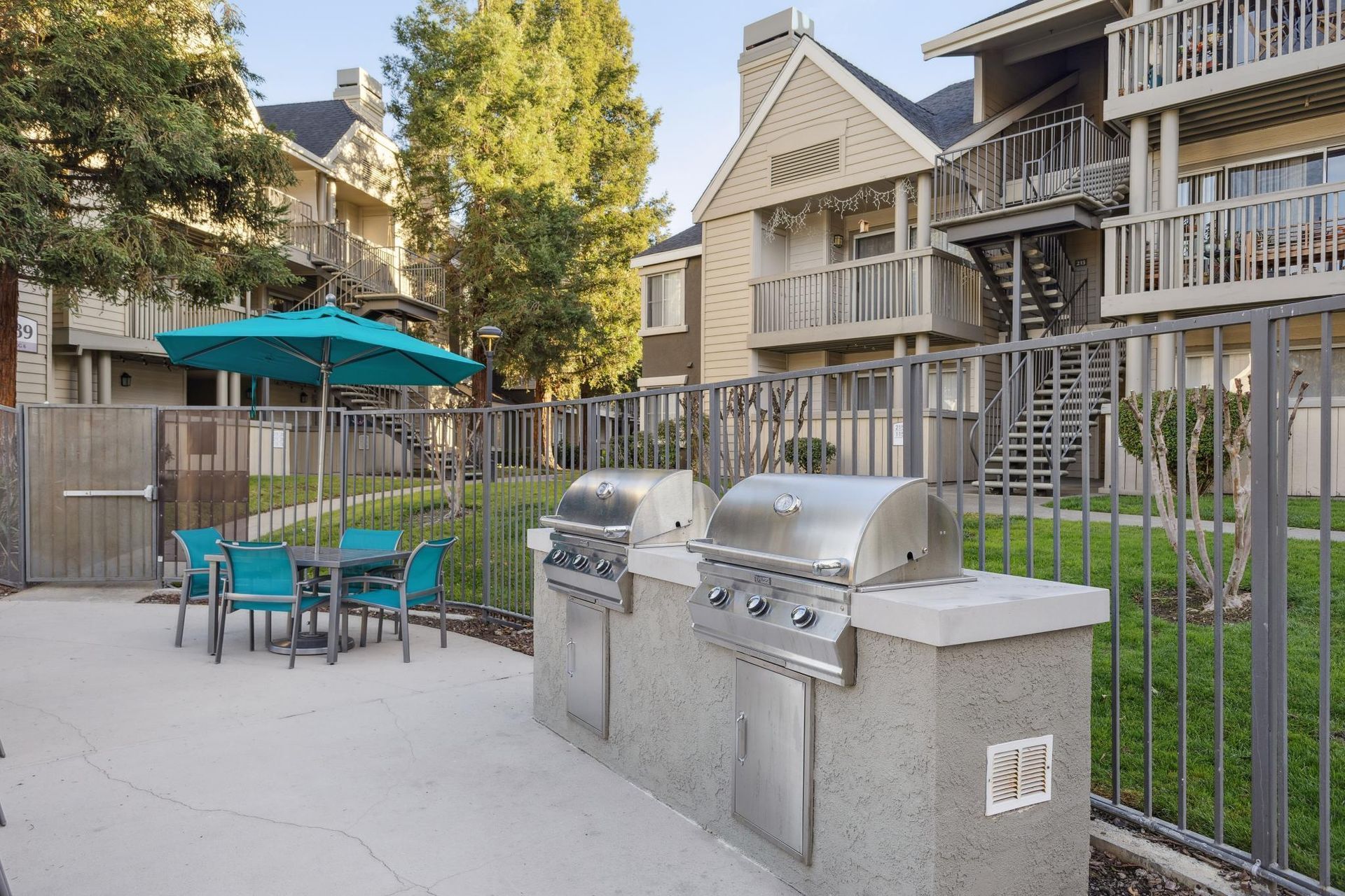 Apartment courtyard with grills, teal patio table and chairs, and a green umbrella near a fence