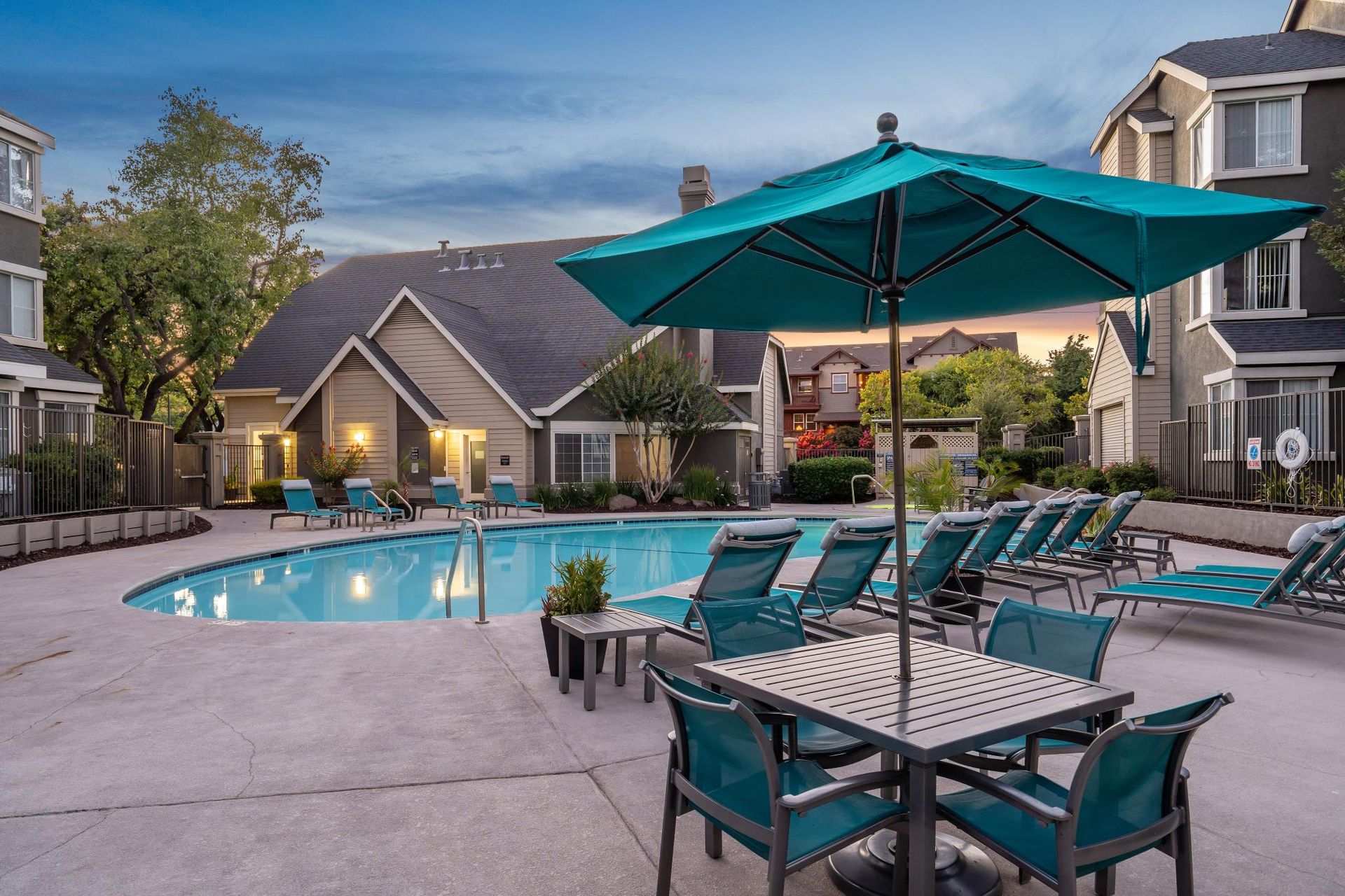 Apartment courtyard with pool, lounge chairs, and teal umbrellas at dusk