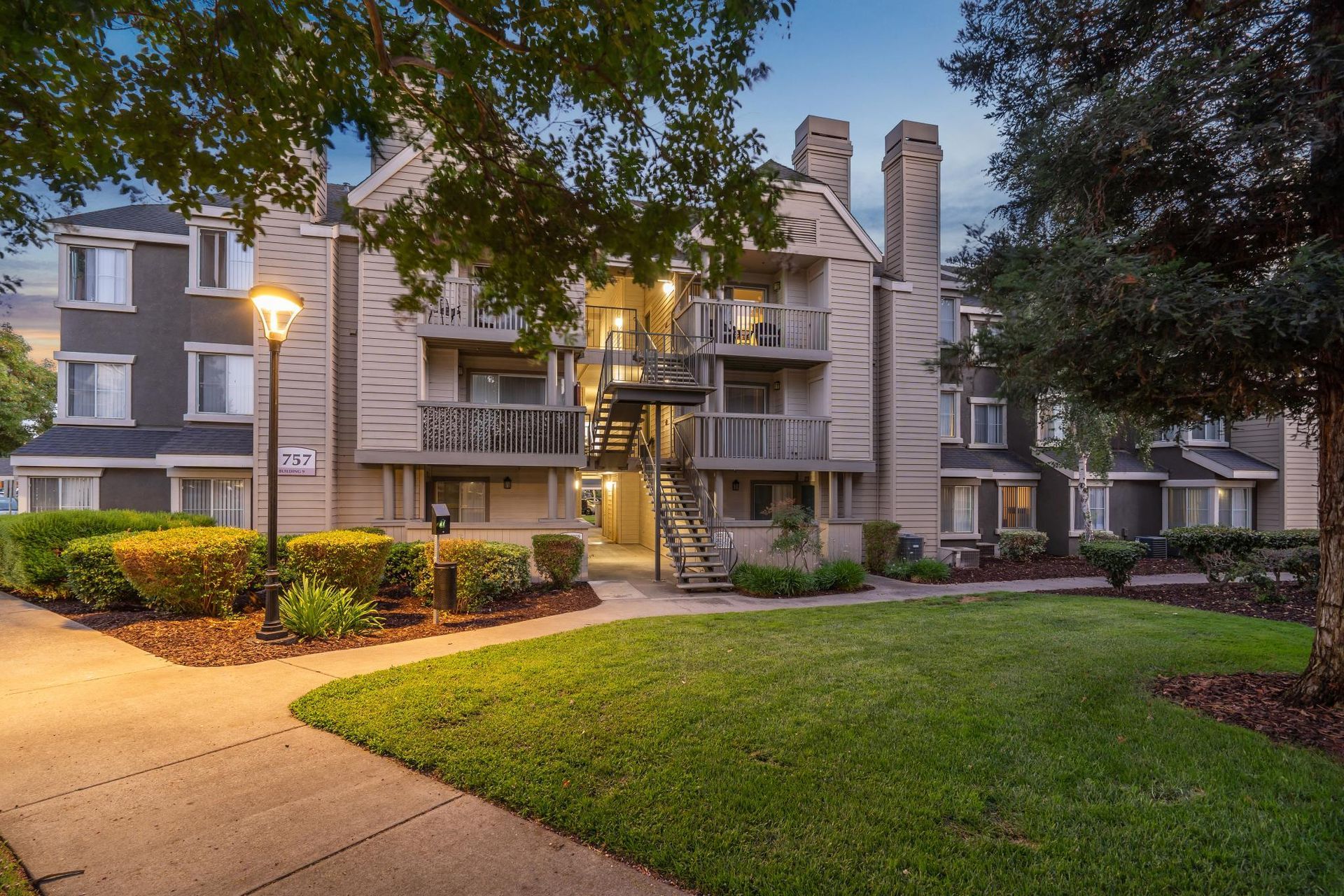 Apartment complex courtyard at dusk with lit walkway, balconies, trees, and landscaped lawn