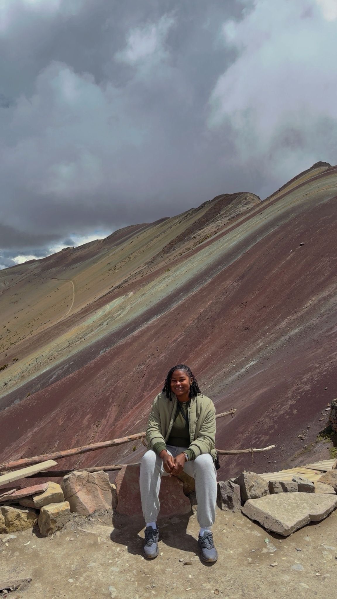 A person sits on a low rock wall in front of a dramatic, multi-colored layered mountain slope under a cloudy sky.