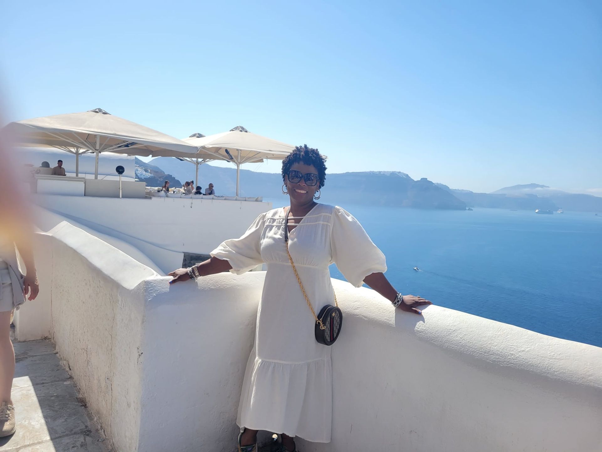 A person in a white midi dress stands on a balcony overlooking the deep blue sea and islands in Santorini, Greece.