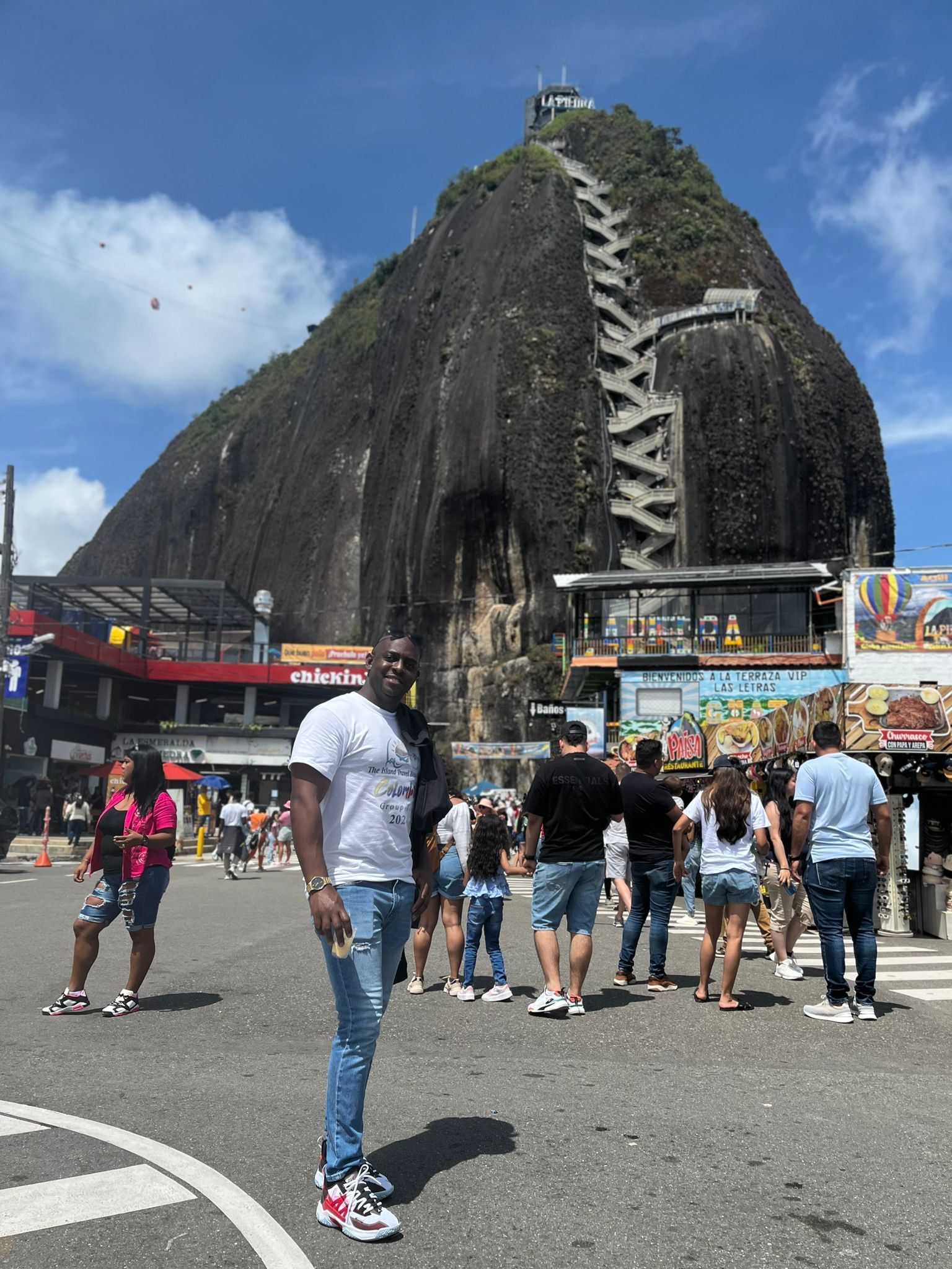A person stands in a plaza before El Peñón de Guatapé, a large monolith in Colombia with a staircase built into its side.