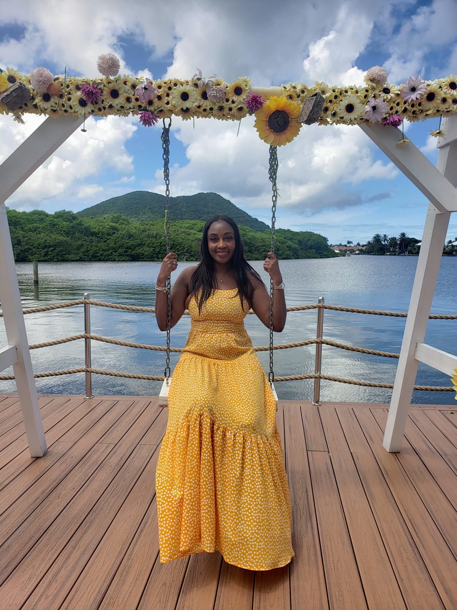 A person in a yellow floral dress sitting on a swing decorated with sunflowers, set on a wooden deck by a lake and hills.