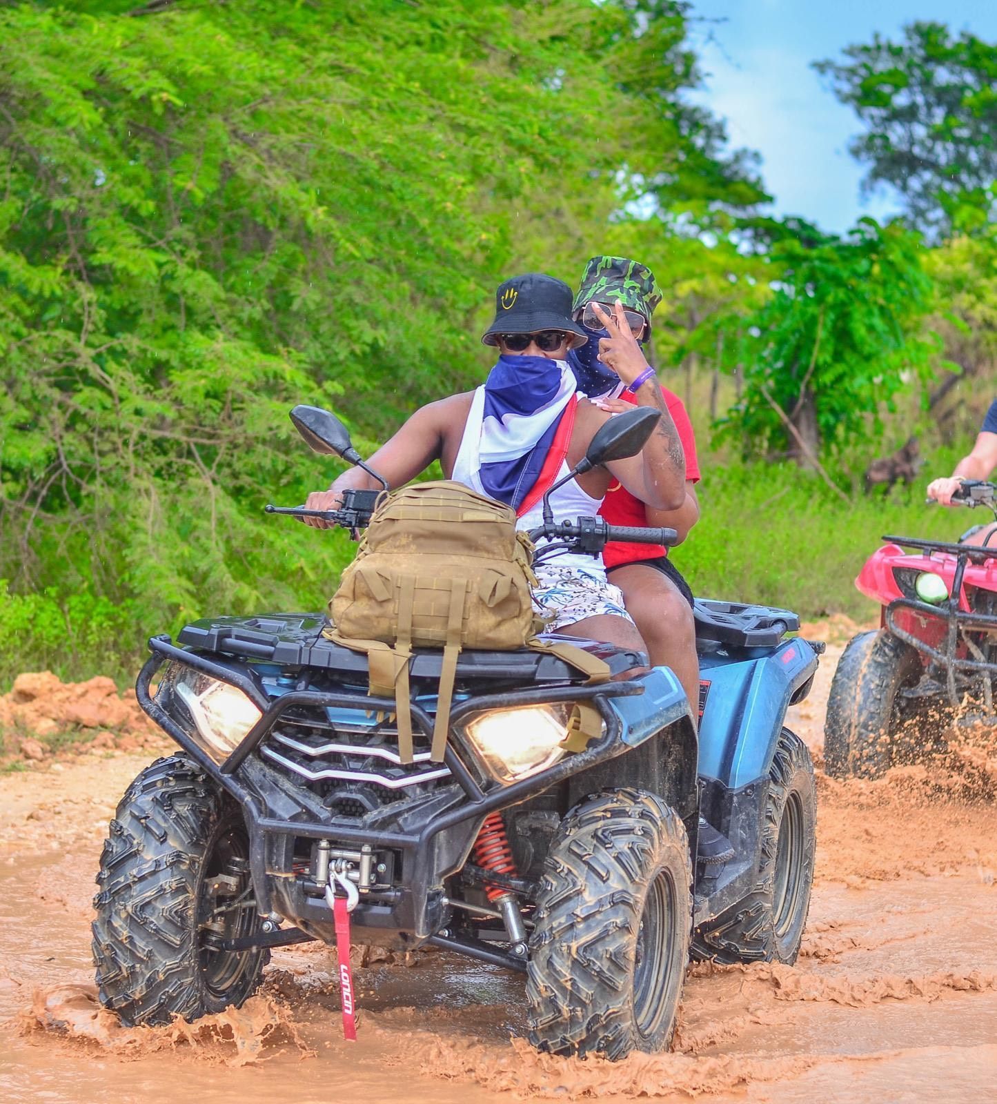 Two people riding a blue ATV through a muddy, outdoor trail, with a second ATV visible in the background.