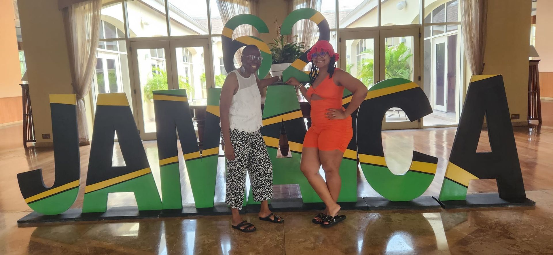 Two people stand smiling between large letters spelling JAMAICA in green, black, and gold colors inside a bright lobby.