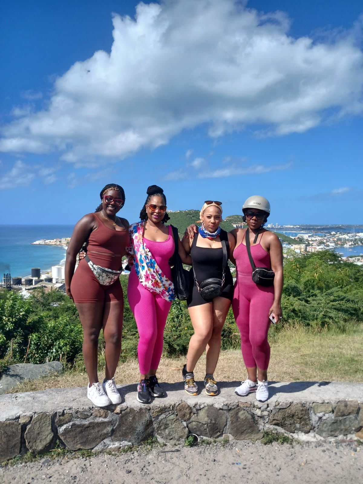 Four friends in matching pink and brown outfits stand smiling before a scenic tropical coastal overlook under a blue sky.