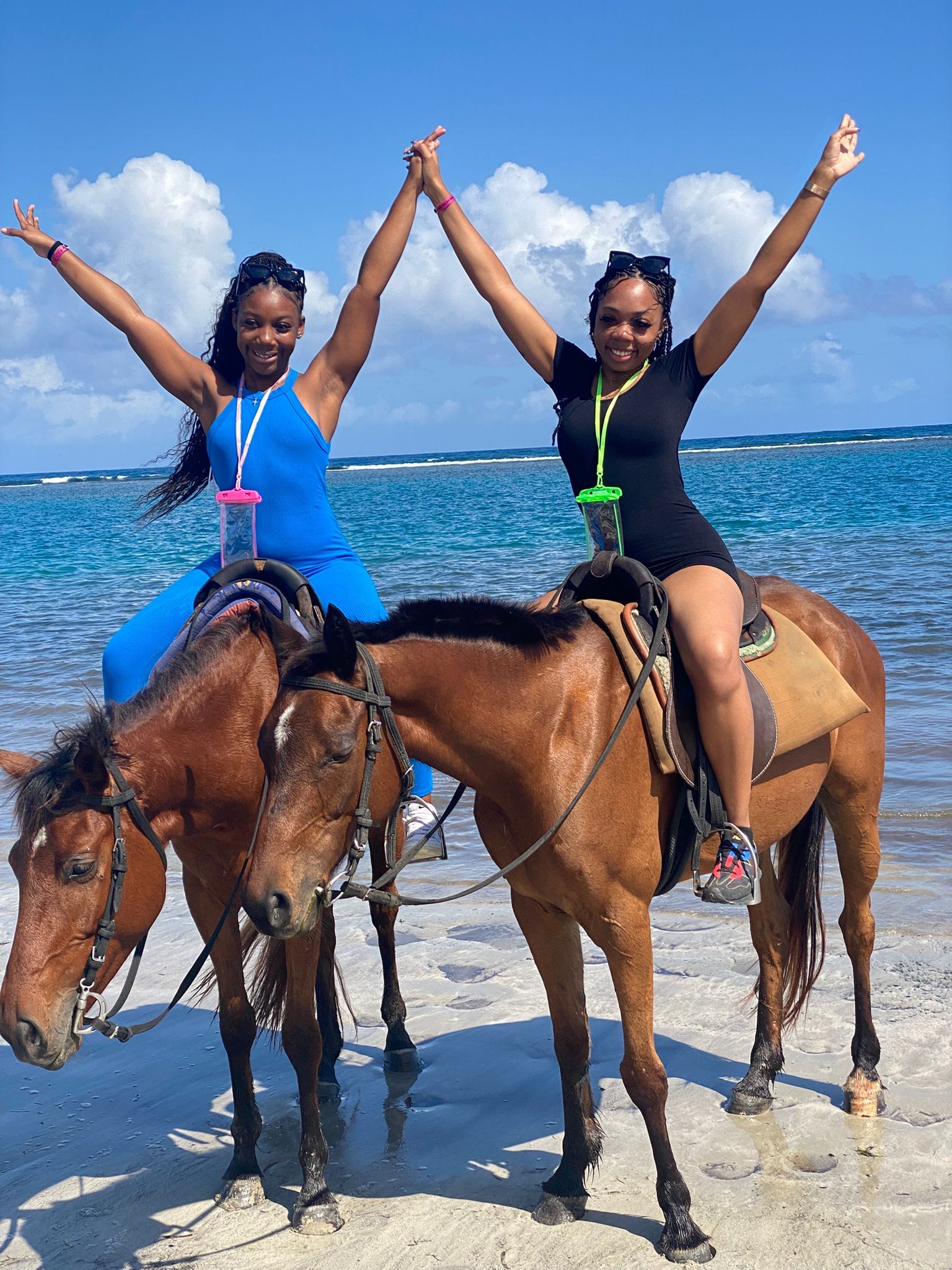 Two people with arms raised in joy, riding horses on a sunny beach with blue water and sky in the background.
