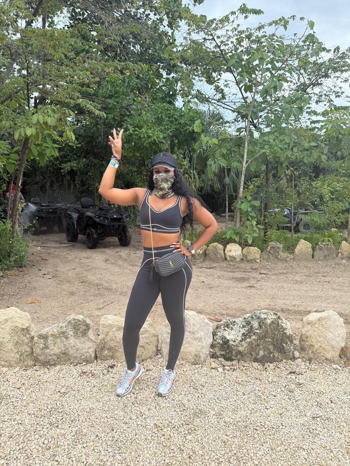A person in a black athletic set and cap poses with a peace sign outdoors near gravel, rocks, and lush green trees.