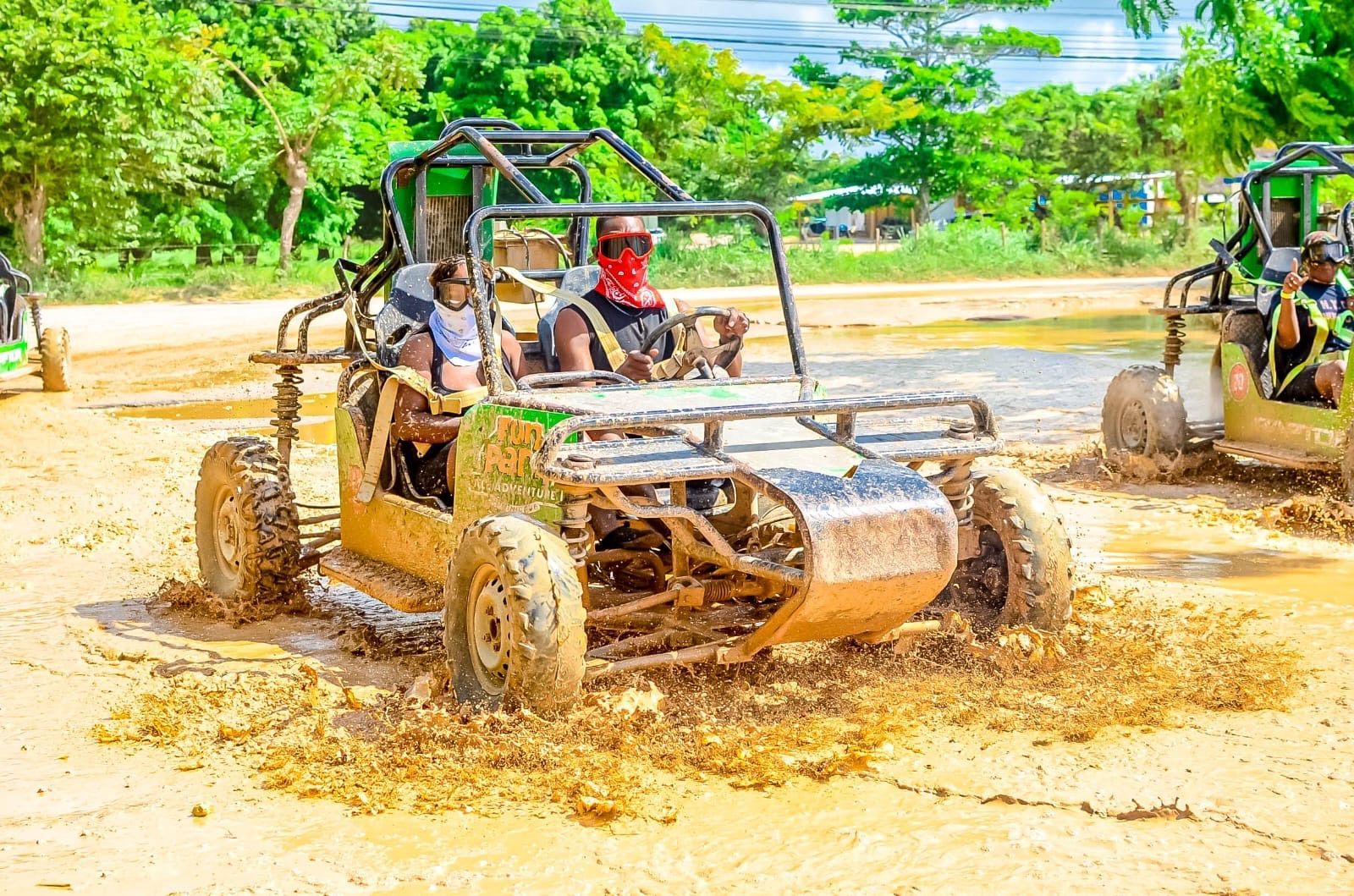 People driving a dune buggy through a muddy, outdoor trail on a sunny day.