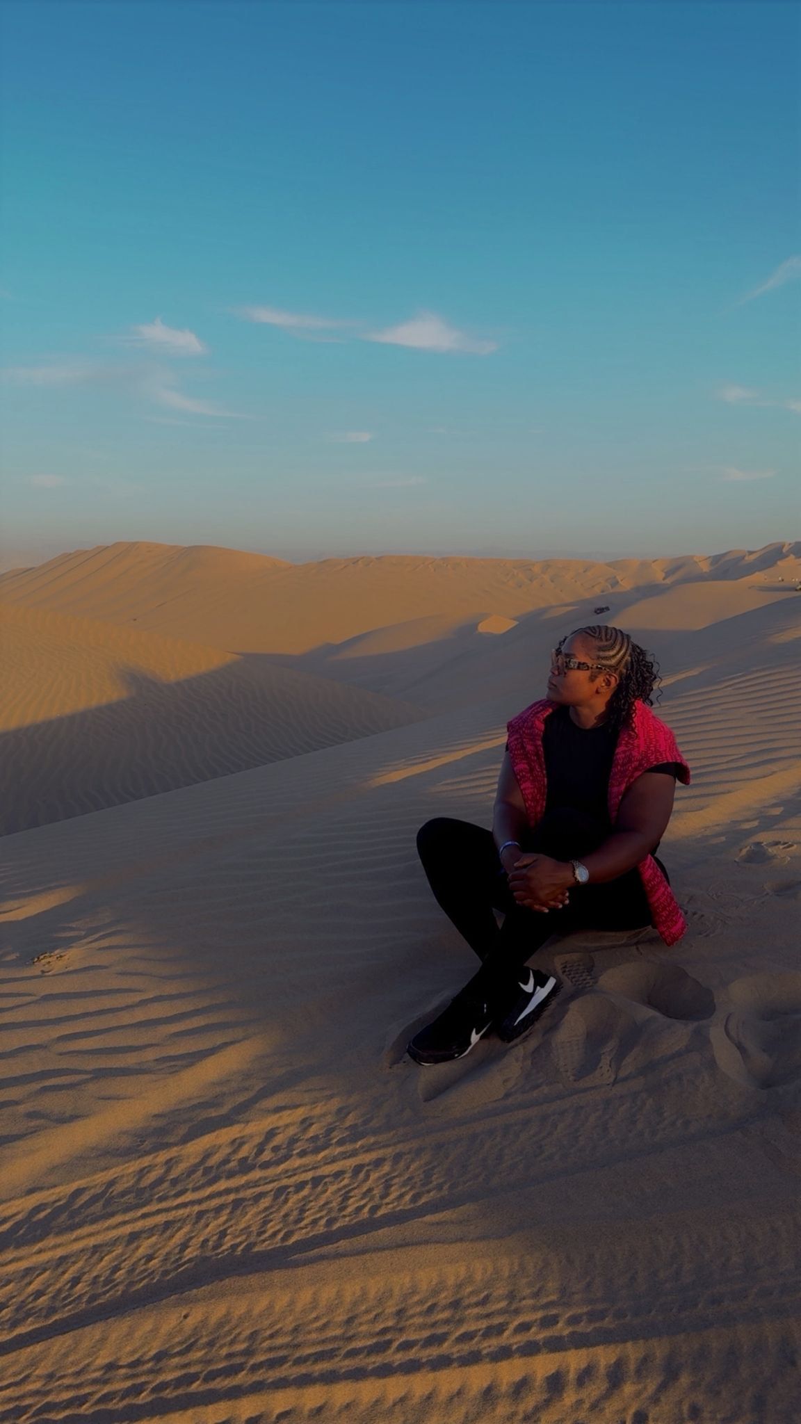 A person in a red jacket sitting on sand dunes in a desert landscape under a clear blue sky.
