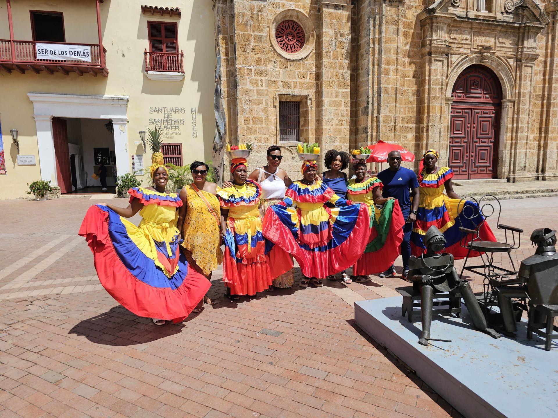A group in traditional Colombian-flag-colored dresses poses in a historic plaza with stone buildings and a statue.