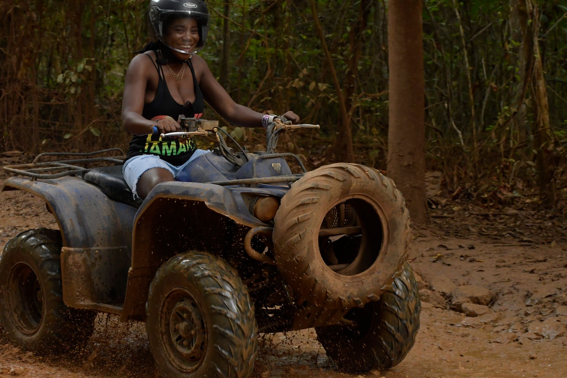 A person smiling while wearing a helmet and riding a muddy ATV through a forest.