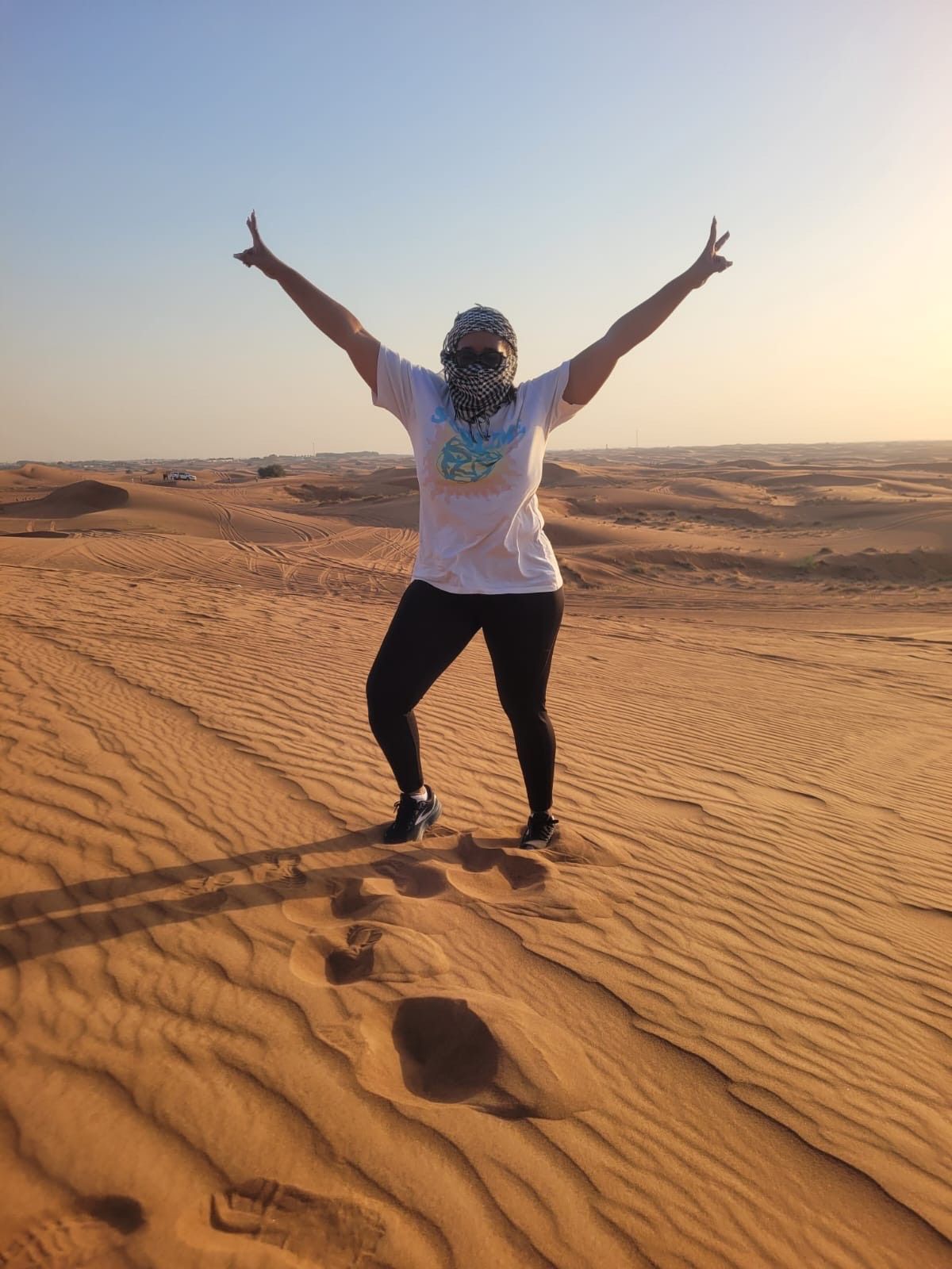 A person wearing a white t-shirt and black leggings stands in a desert with arms raised against a clear sky.