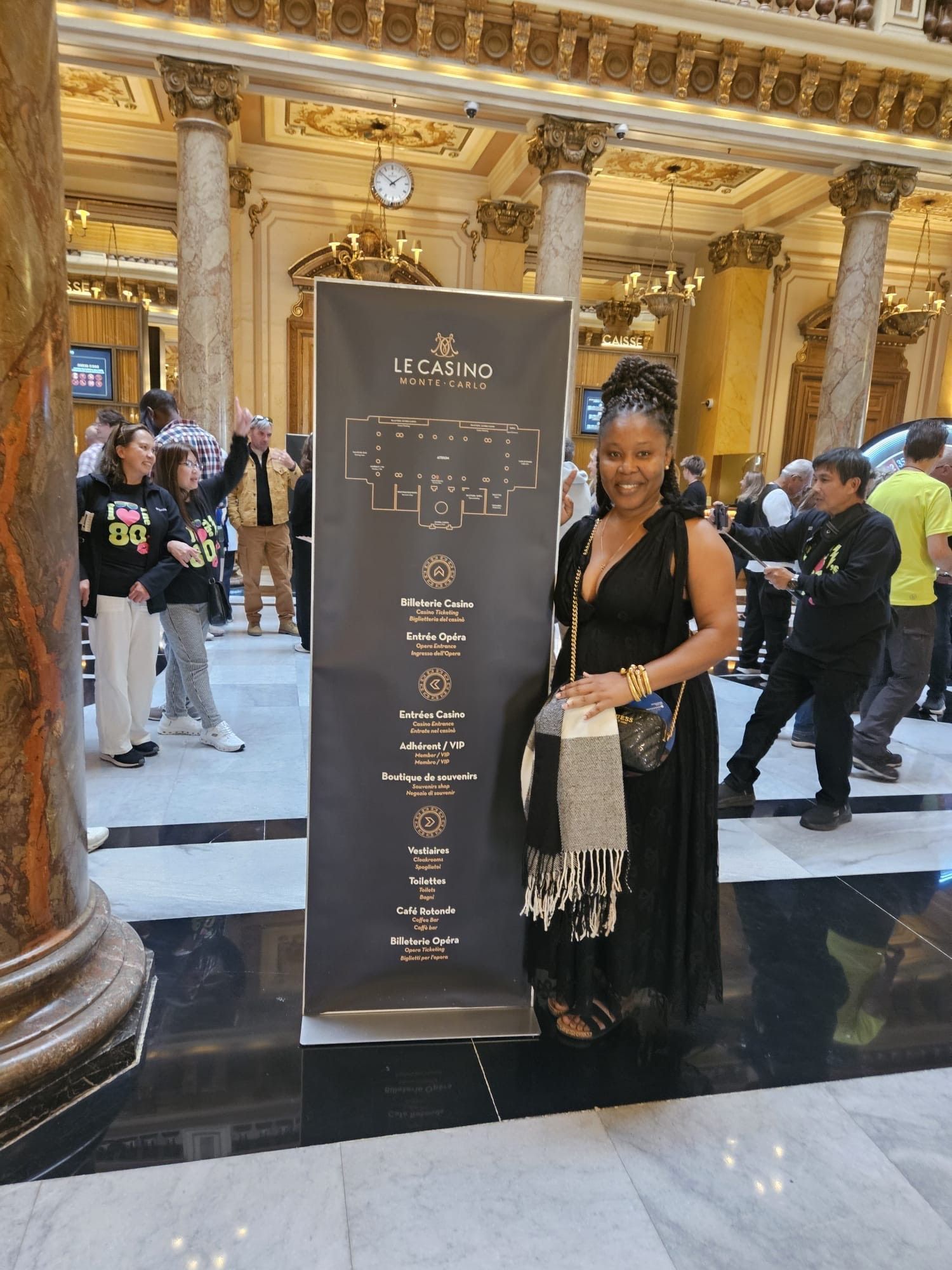A person in a black dress stands smiling next to a tall event information sign inside an ornate, marble-columned hall.