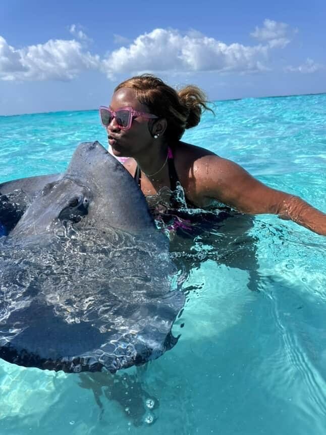 A woman in sunglasses puckers her lips to kiss a large stingray in clear turquoise ocean water.