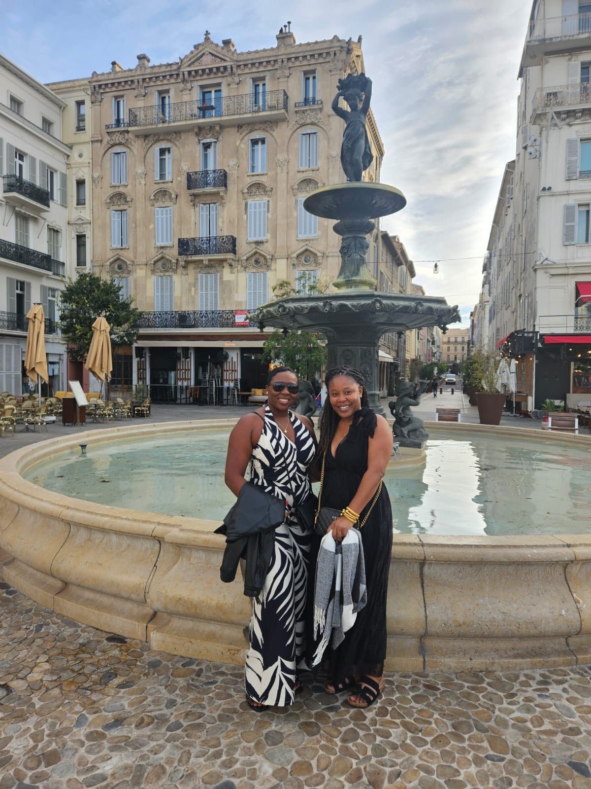 Two people stand smiling in front of a historic stone fountain in a sunny, European-style city square.