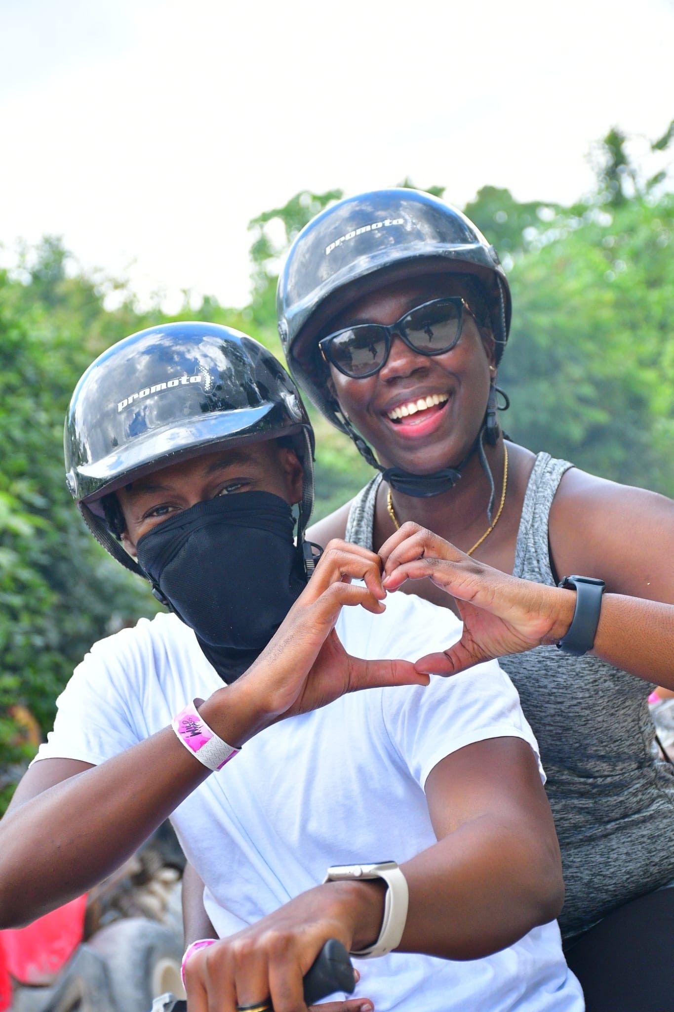 Two people wearing helmets and sunglasses on a motorcycle make a heart shape with their hands while smiling outdoors.