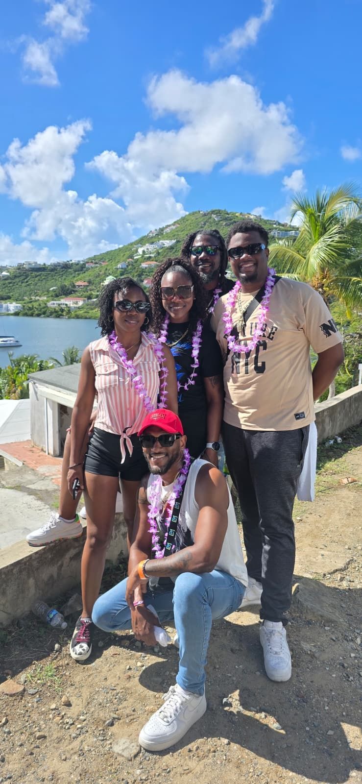 Five people wearing floral leis pose for a photo with a tropical harbor and hilly landscape in the background.