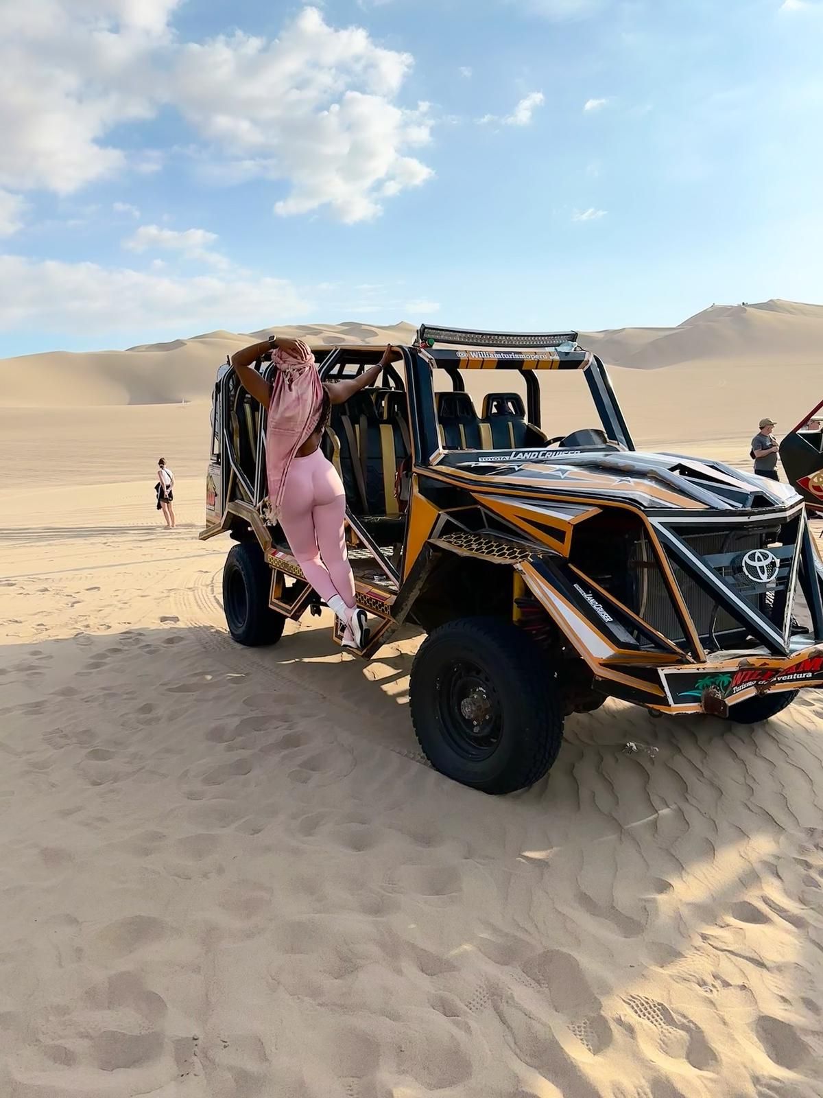 A person in pink clothing stands on the side of a large, yellow, open-air dune buggy in a vast sandy desert.