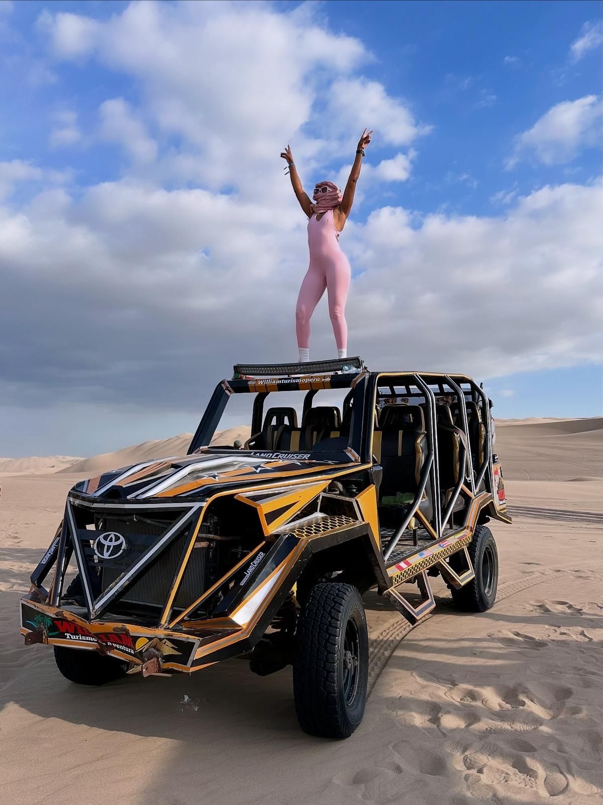 A person in a pink jumpsuit stands with arms raised atop a gold and black off-road vehicle in a sandy desert.