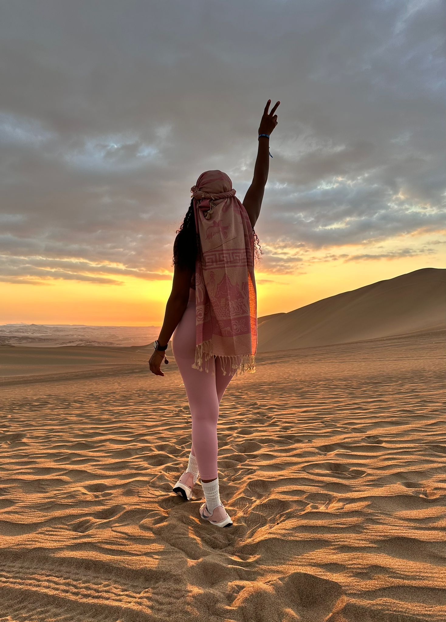 A person stands in a sandy desert at sunset, wearing light leggings and a scarf, with one arm raised toward the sky.