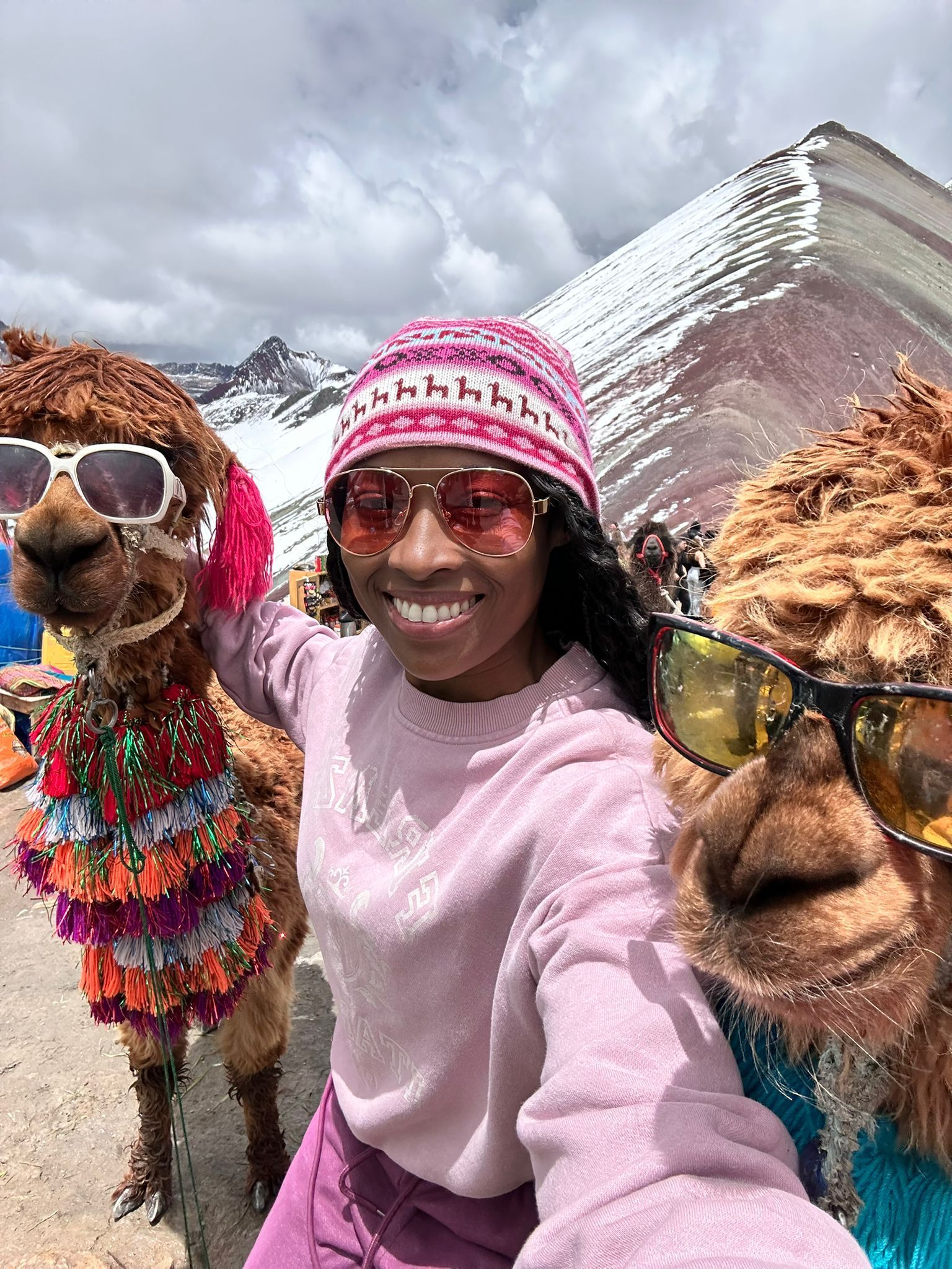 A smiling person in a pink sweater and knit cap takes a selfie with two alpacas wearing sunglasses near a snowy mountain.