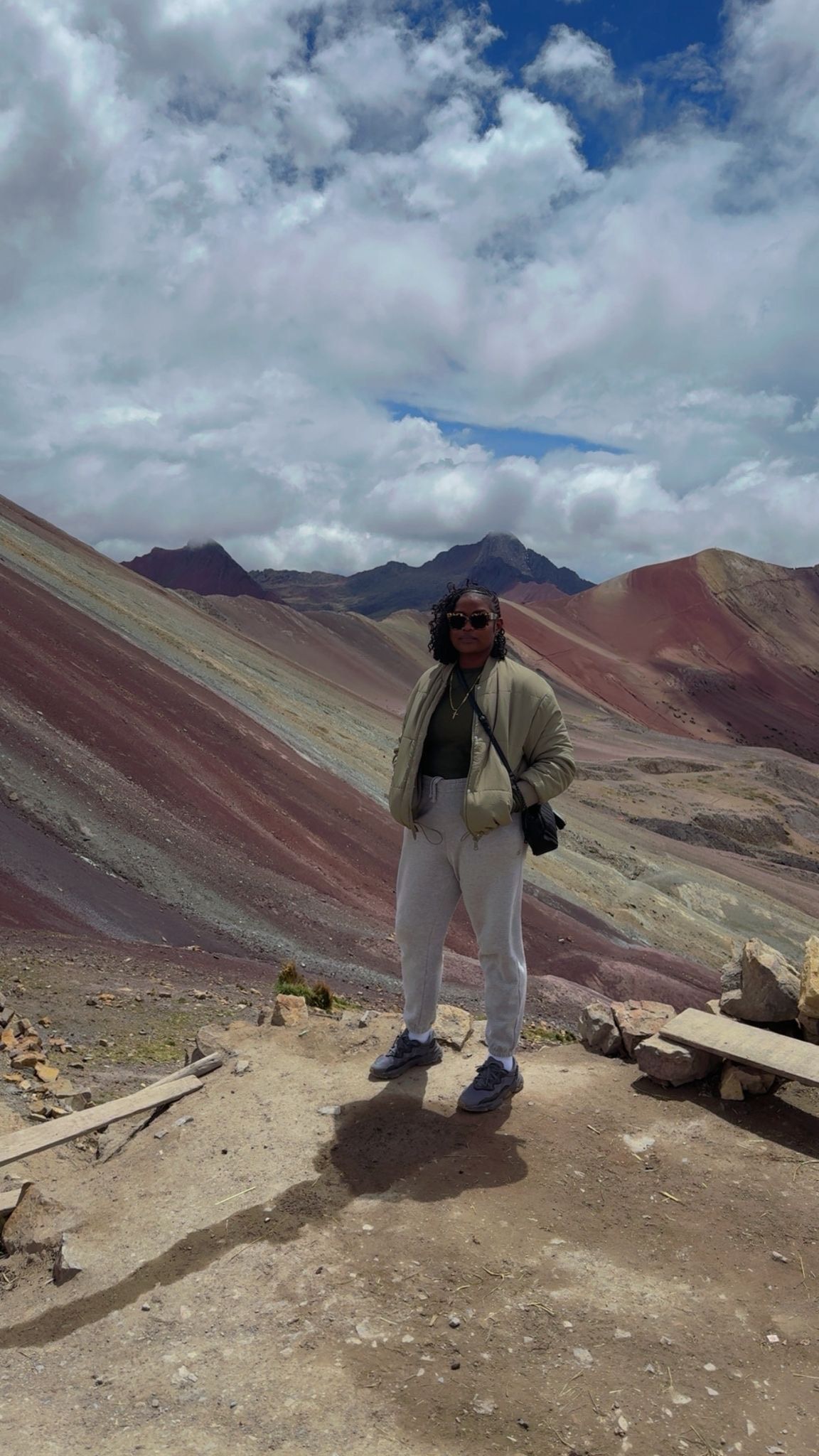 A person in a light jacket and pants standing on a rocky trail with the colorful, striped slopes of Vinicunca, Peru.