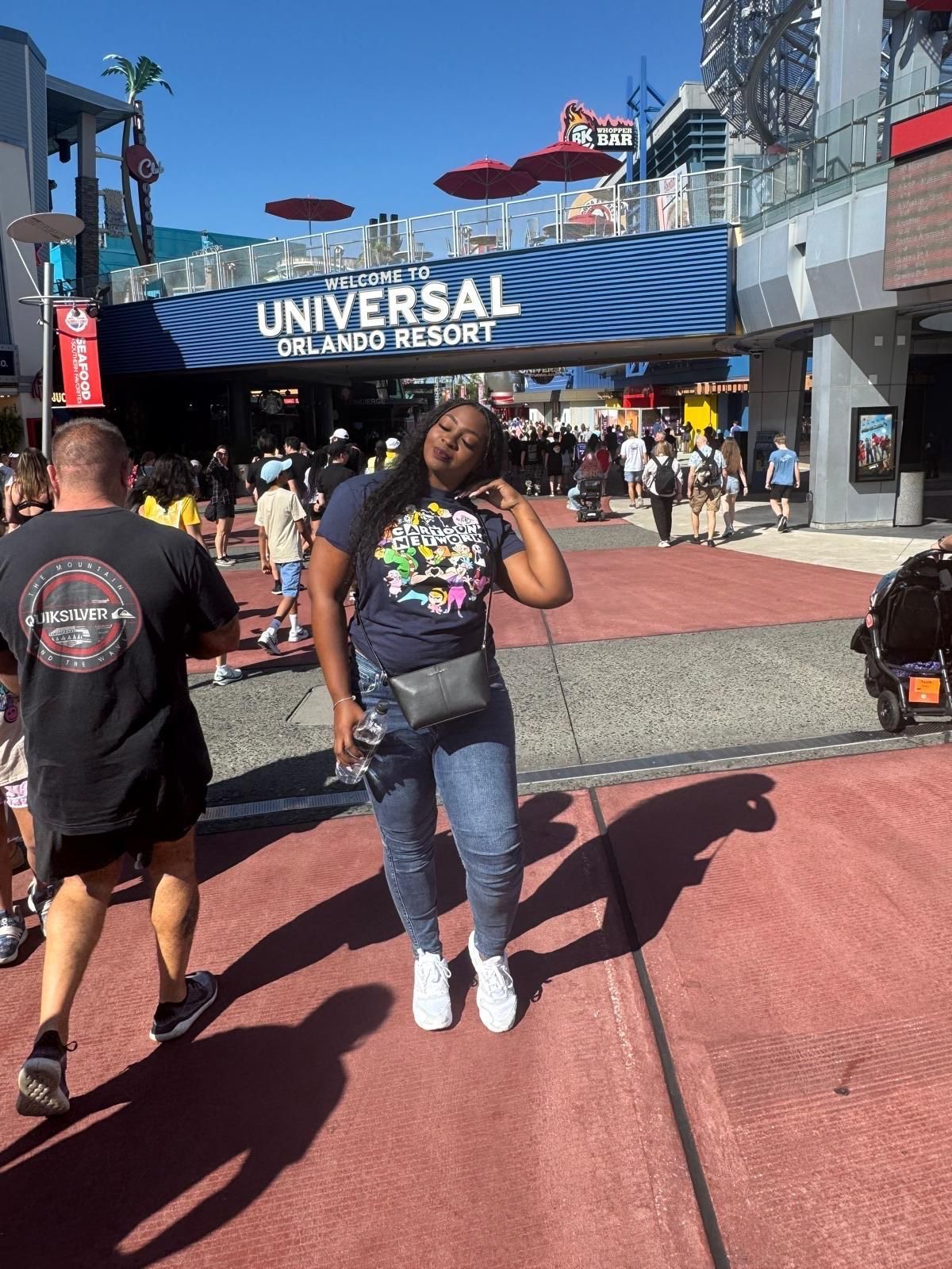 A person poses in front of the Universal Orlando Resort entrance under a bright blue sky with a blue overhead sign.