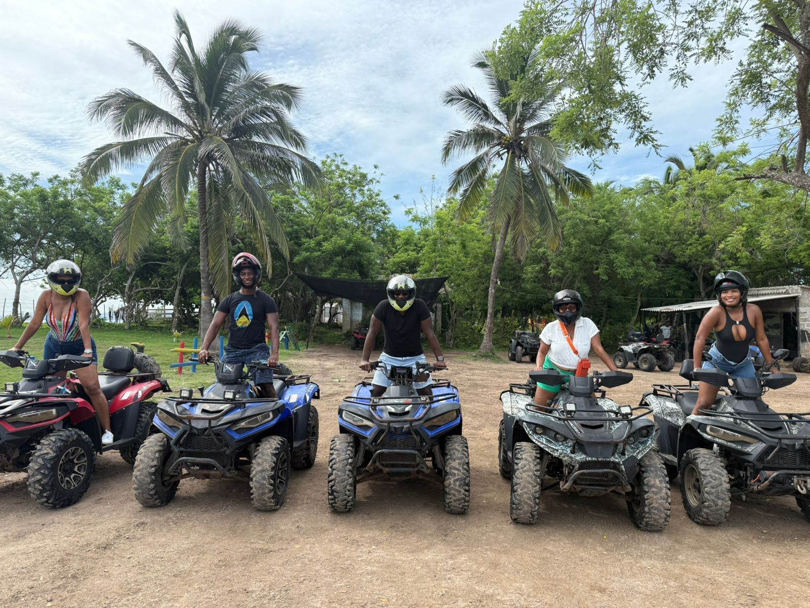 Five people on ATVs posing for a photo on a dirt path, with palm trees and green foliage in the background.