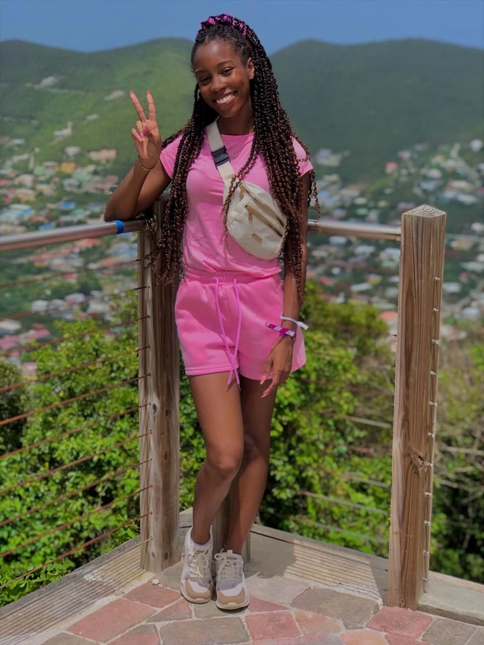 A smiling person wearing a pink outfit and crossbody bag poses with a peace sign, overlooking a hilly landscape.