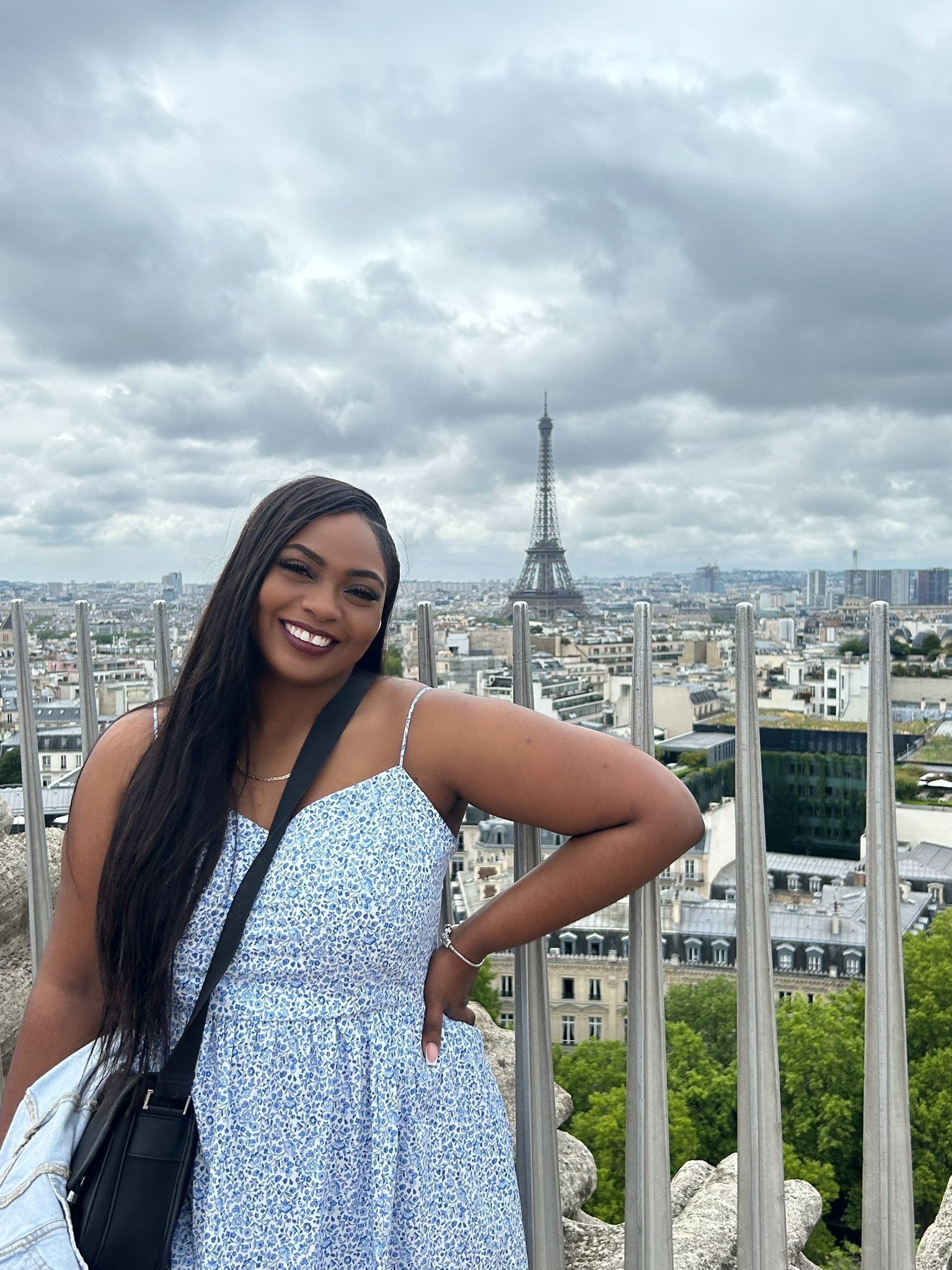 A smiling person stands in front of a metal railing overlooking a cityscape with the Eiffel Tower in the distance.