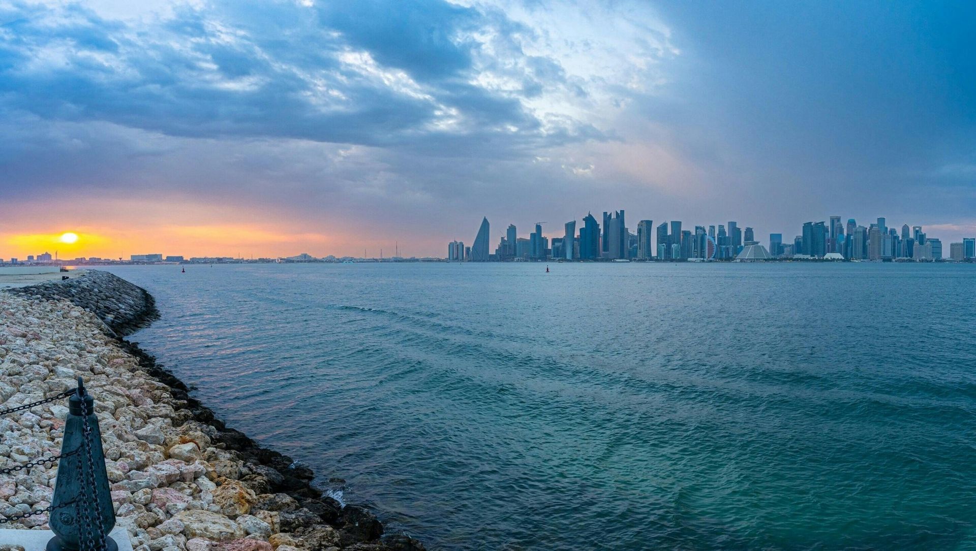 Skyline of Doha, Qatar, at sunset, over water. Cloudy sky, cityscape silhouette, and stone breakwater.