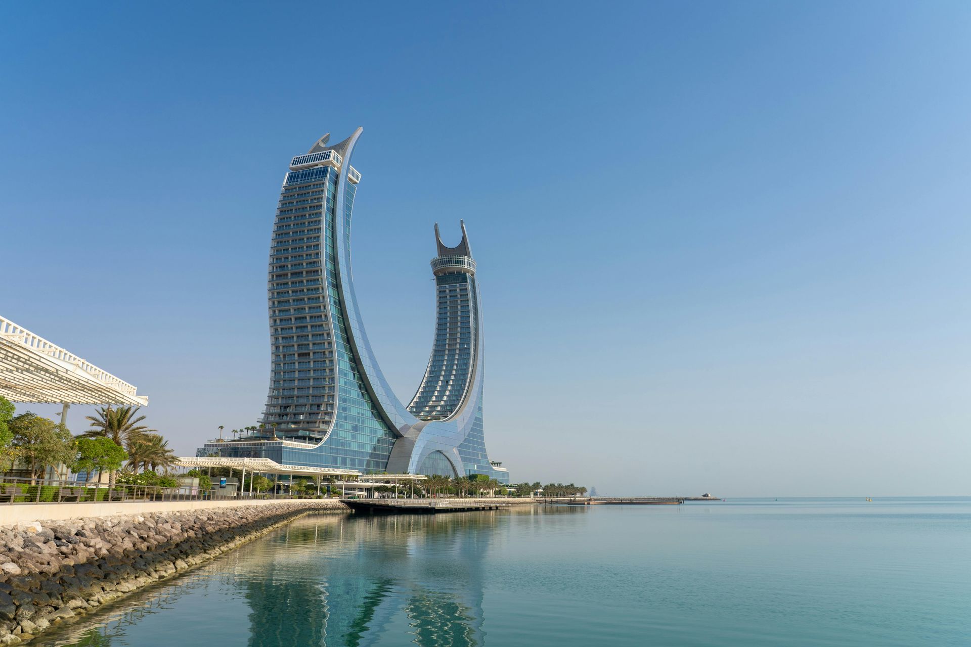 Two tall, arching skyscrapers reflect in calm water under a clear blue sky.