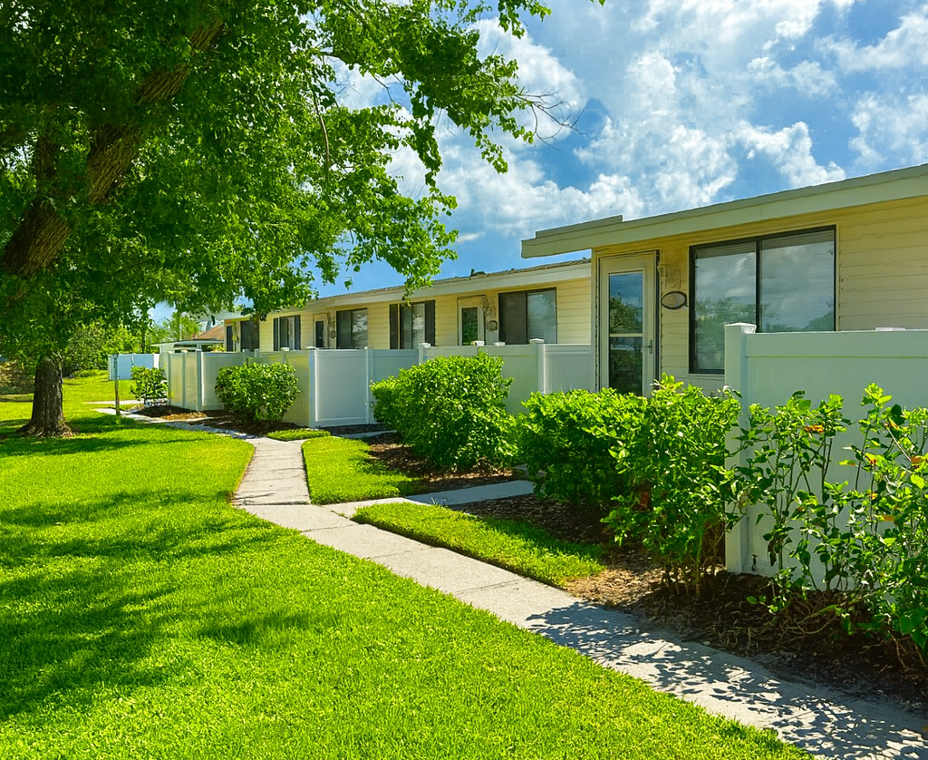 Lawn and sidewalk lead to a row of beige buildings with white fences, under a blue sky.