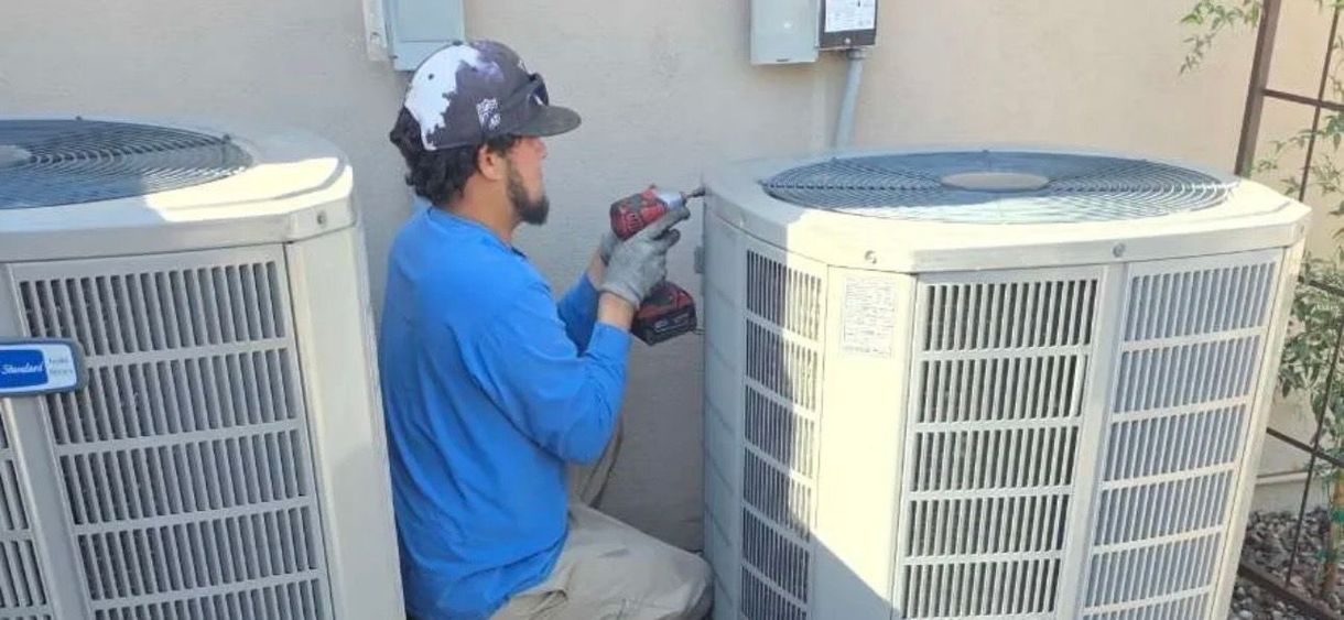 Technician kneels between two outdoor HVAC units, using a drill beside a building wall.