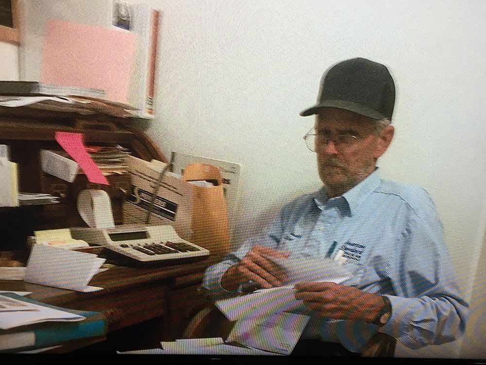 Man sitting at a desk in an office, holding papers and looking toward the camera.