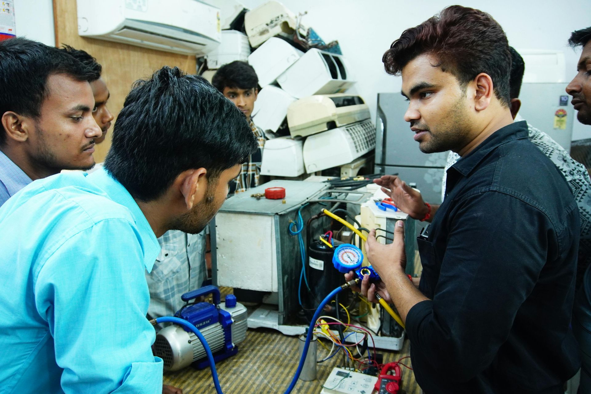 Three men inspecting wiring on a cluttered workbench with a blue motor in a workshop