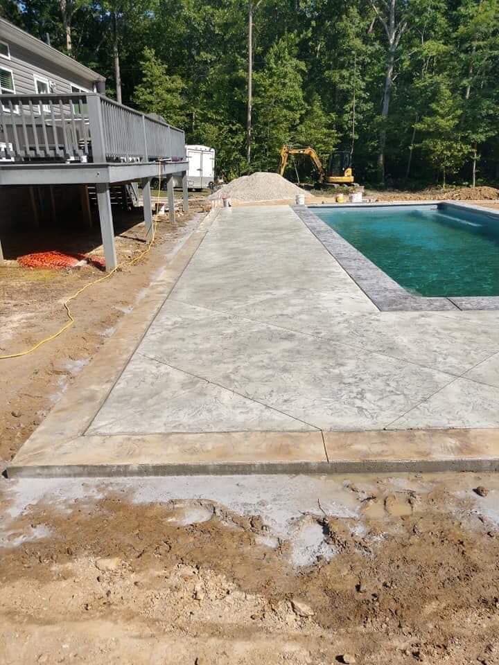 Concrete patio beside a pool under construction, with a deck on the left and trees in the background