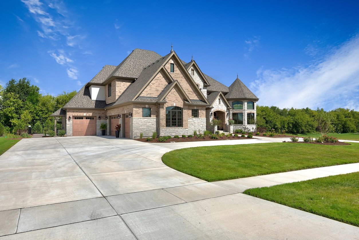 Large stone suburban house with a three-car garage, wide driveway, and manicured lawn under a blue sky