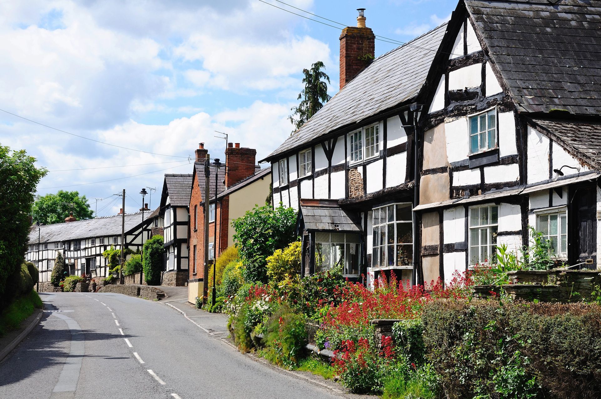 A black and white historic listed Tudor period house lines the road in Herefordshire, England. Beautiful flowers are shown in the garden.