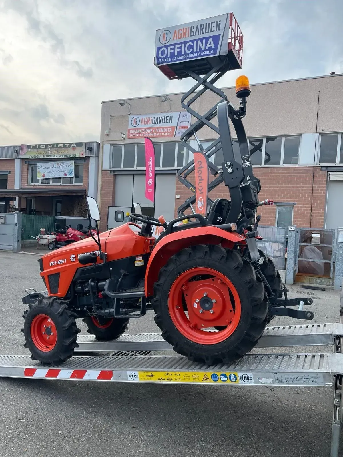 Trattore rosso con braccio escavatore posteriore sollevato su un rimorchio davanti a un edificio, sotto un cielo nuvoloso.