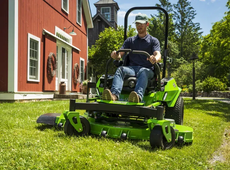 Un uomo alla guida di un tosaerba verde in un giardino soleggiato accanto a una casa rossa.
