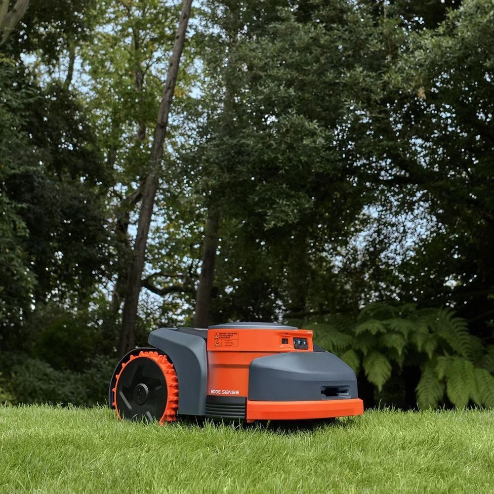 Robot tagliaerba arancione e grigio sull'erba in un cortile alberato.