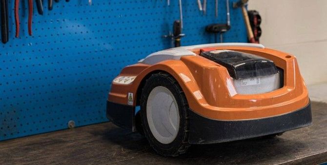 Un robot tagliaerba arancione su un banco da lavoro davanti a una parete con pannelli forati blu.